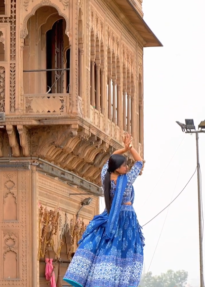 Woman in a blue traditional outfit standing in front of an ornate wooden building.